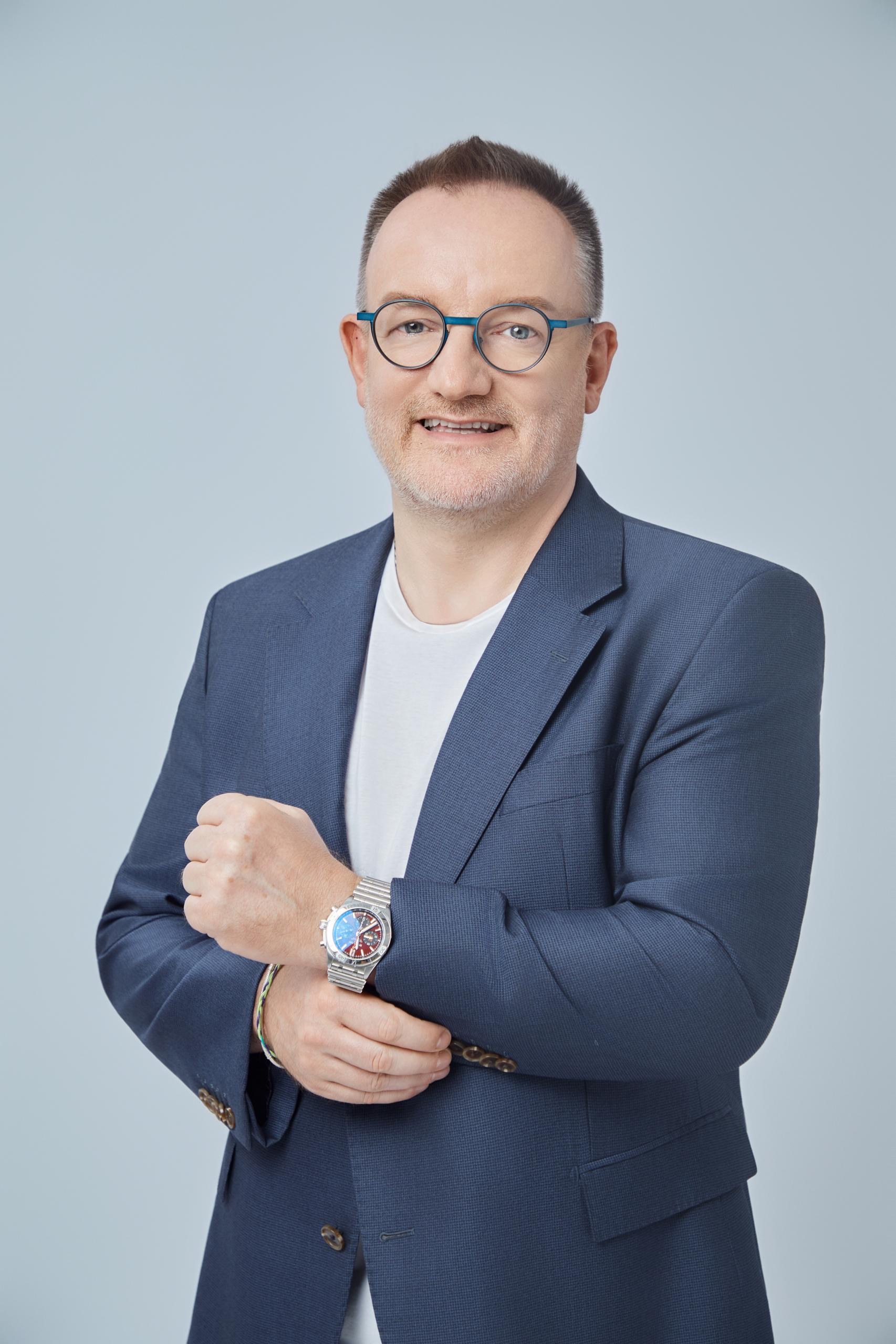 Ein Mann mit Brille lächelt freundlich, trägt ein blaues Sakko und zeigt seine Uhr